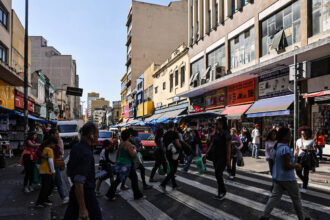 A imagem mostra um cruzamento movimentado em uma área urbana, com várias pessoas atravessando a rua em uma faixa de pedestres. Ao fundo, há prédios de diferentes alturas e estilos arquitetônicos, além de lojas com toldos coloridos. Veículos estão estacionados e em movimento na rua, e o céu está claro, indicando um dia ensolarado.