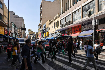 Os nomes e sobrenomes mais comuns no Brasil, segundo o Censo A imagem mostra um cruzamento movimentado em uma área urbana, com várias pessoas atravessando a rua em uma faixa de pedestres. Ao fundo, há prédios de diferentes alturas e estilos arquitetônicos, além de lojas com toldos coloridos. Veículos estão estacionados e em movimento na rua, e o céu está claro, indicando um dia ensolarado.