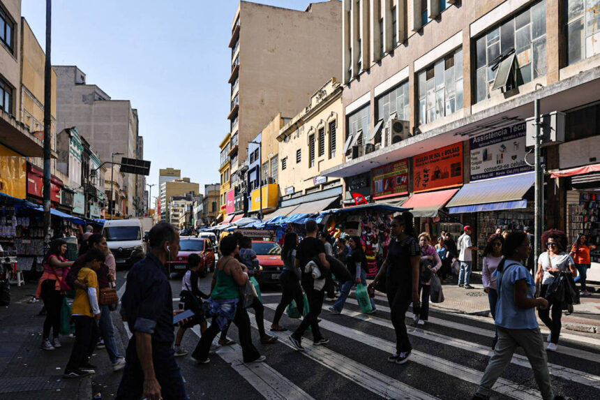 A imagem mostra um cruzamento movimentado em uma área urbana, com várias pessoas atravessando a rua em uma faixa de pedestres. Ao fundo, há prédios de diferentes alturas e estilos arquitetônicos, além de lojas com toldos coloridos. Veículos estão estacionados e em movimento na rua, e o céu está claro, indicando um dia ensolarado.