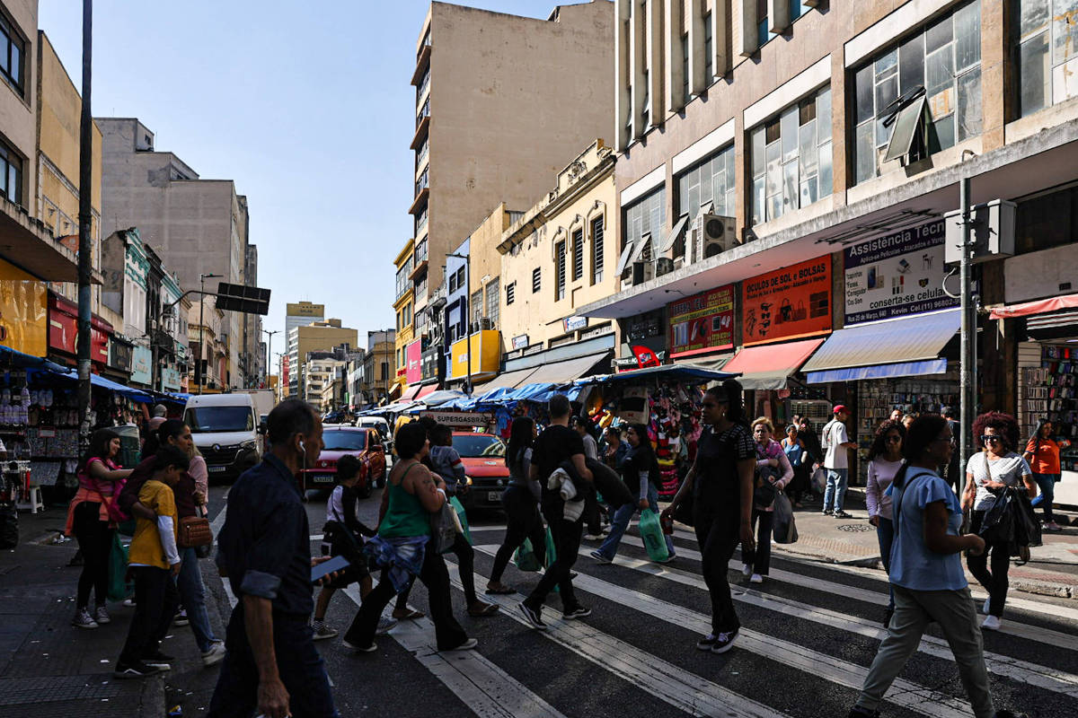 Os nomes e sobrenomes mais comuns no Brasil, segundo o Censo A imagem mostra um cruzamento movimentado em uma área urbana, com várias pessoas atravessando a rua em uma faixa de pedestres. Ao fundo, há prédios de diferentes alturas e estilos arquitetônicos, além de lojas com toldos coloridos. Veículos estão estacionados e em movimento na rua, e o céu está claro, indicando um dia ensolarado.