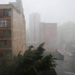 Calor em SP na sexta e sábado antes da tempestade A imagem mostra um cenário urbano durante uma forte chuva. Vários prédios são visíveis, com um deles em primeiro plano apresentando janelas e uma fachada de tijolos. A visibilidade é baixa devido à intensidade da chuva, e árvores balançam com o vento. O ambiente parece molhado e cinzento, refletindo as condições climáticas adversas.