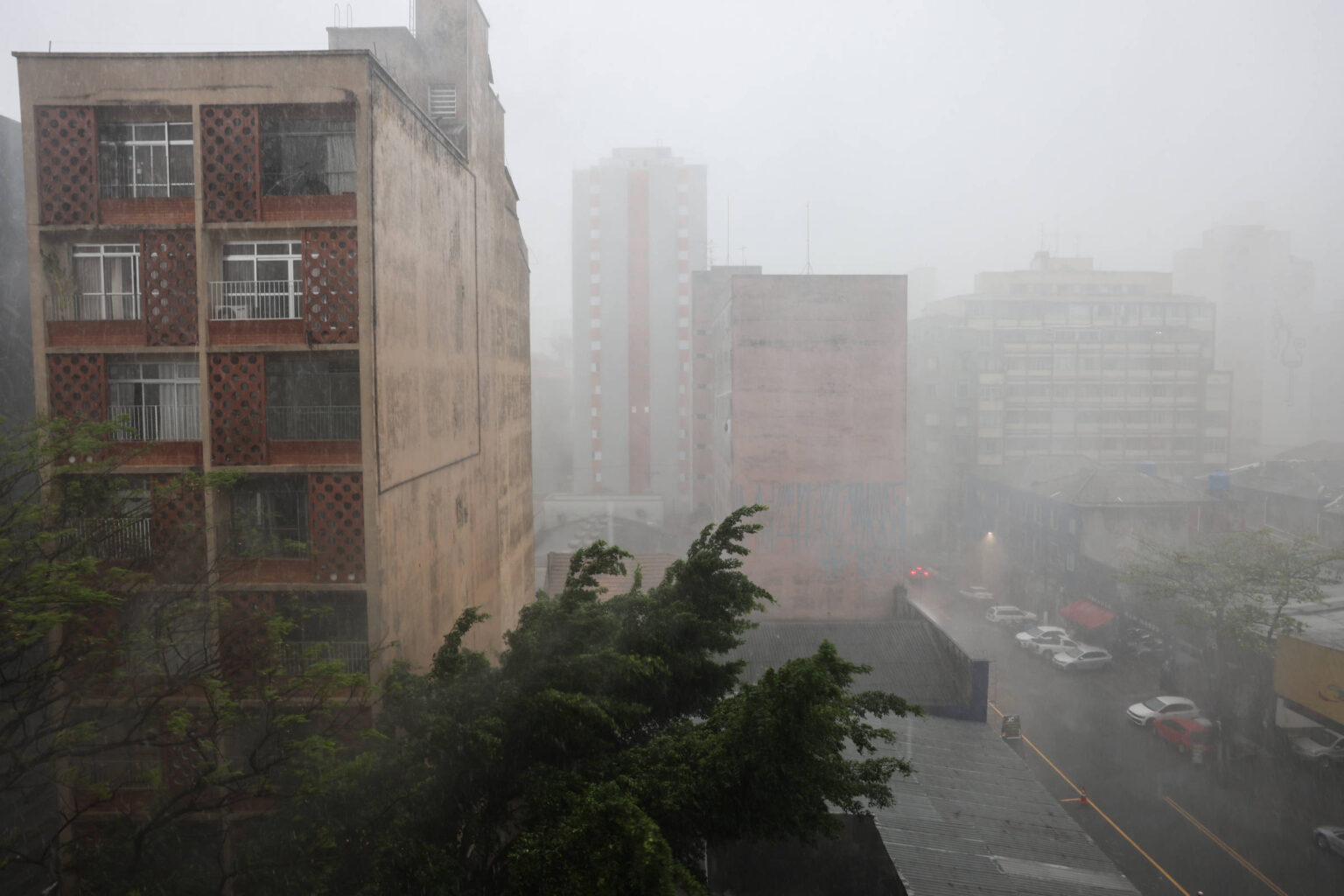 Calor em SP na sexta e sábado antes da tempestade A imagem mostra um cenário urbano durante uma forte chuva. Vários prédios são visíveis, com um deles em primeiro plano apresentando janelas e uma fachada de tijolos. A visibilidade é baixa devido à intensidade da chuva, e árvores balançam com o vento. O ambiente parece molhado e cinzento, refletindo as condições climáticas adversas.
