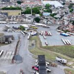 Terminal de ônibus com vários veículos estacionados em áreas delimitadas, próximo a uma região urbana densa com muitas construções e casas. Estradas internas conectam as áreas de estacionamento e há vegetação ao redor do terminal.