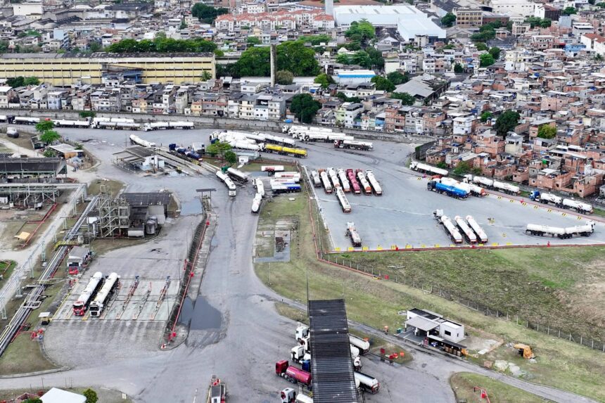 Terminal de ônibus com vários veículos estacionados em áreas delimitadas, próximo a uma região urbana densa com muitas construções e casas. Estradas internas conectam as áreas de estacionamento e há vegetação ao redor do terminal.