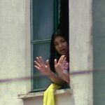 Mulher com cabelo preto aparece na janela aberta de um prédio claro, com as mãos levantadas em sinal de parada ou comunicação. Ela veste roupa clara e há um tecido amarelo pendurado no parapeito da janela.