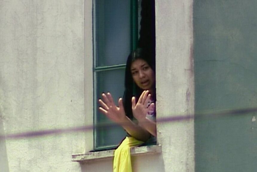 Mulher com cabelo preto aparece na janela aberta de um prédio claro, com as mãos levantadas em sinal de parada ou comunicação. Ela veste roupa clara e há um tecido amarelo pendurado no parapeito da janela.