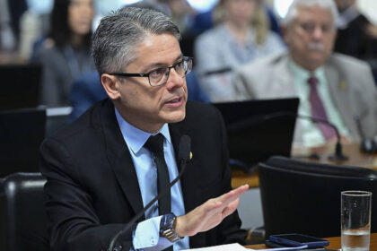 Alessandro Vieira cobra próximo passo do RJ sobre decisões Homem de cabelos grisalhos e óculos, vestido com terno preto e gravata azul, fala em microfone durante audiência. Pessoas desfocadas ao fundo e copo de água na mesa.