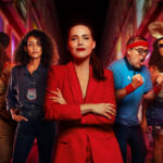 Cinco pessoas em pé em uma rua iluminada à noite. No centro, mulher de terno vermelho com braços cruzados. Ao redor, pessoas com roupas casuais e estilos variados. Texto