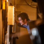 Homem de camiseta azul inclina-se para examinar um painel elétrico aberto em ambiente interno com iluminação amarelada. O foco está no rosto concentrado e na mão que toca o painel.