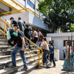 Grupo de pessoas subindo escada externa com corrimão amarelo em prédio branco. Ambiente ao ar livre com árvores e sol forte. Algumas pessoas carregam mochilas e bolsas.