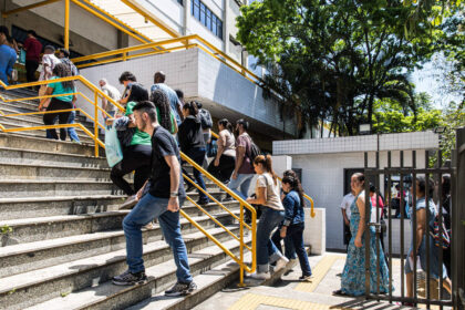 Grupo de pessoas subindo escada externa com corrimão amarelo em prédio branco. Ambiente ao ar livre com árvores e sol forte. Algumas pessoas carregam mochilas e bolsas.