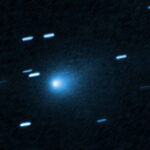 Cometa azul brilhante no centro da imagem com uma cauda difusa ao redor. Traços de estrelas aparecem como linhas claras ao redor, indicando movimento ou longa exposição.