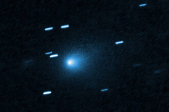 Cometa azul brilhante no centro da imagem com uma cauda difusa ao redor. Traços de estrelas aparecem como linhas claras ao redor, indicando movimento ou longa exposição.