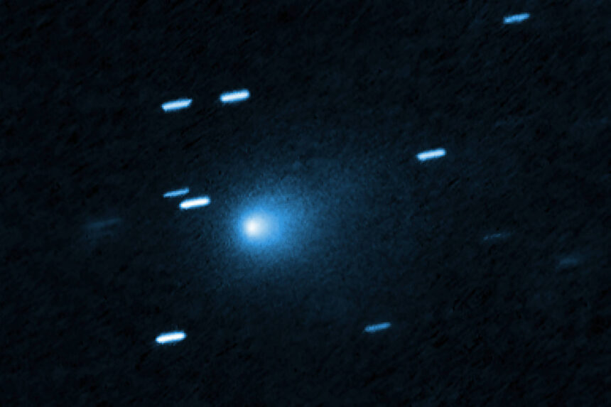 Cometa azul brilhante no centro da imagem com uma cauda difusa ao redor. Traços de estrelas aparecem como linhas claras ao redor, indicando movimento ou longa exposição.