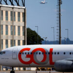Gol registra lucro no 3º trimestre e atualiza projeções Avião da Gol estacionado em primeiro plano próximo a um prédio de janelas retangulares. Ao fundo, outro avião está em aproximação para pouso sob céu azul.