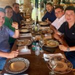 Aliado de Ciro Nogueira viaja com investigados do PCC Seis homens sentados ao redor de uma mesa de madeira em restaurante com decoração rústica, incluindo pilares de tijolos e teto com estrutura de madeira. A mesa está posta com pratos, talheres, copos e garrafas de água. Ao fundo, outras pessoas estão em pé e sentadas, com luz natural entrando pelas janelas.