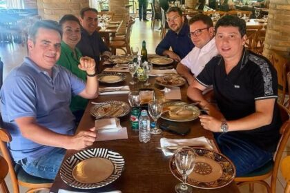 Aliado de Ciro Nogueira viaja com investigados do PCC Seis homens sentados ao redor de uma mesa de madeira em restaurante com decoração rústica, incluindo pilares de tijolos e teto com estrutura de madeira. A mesa está posta com pratos, talheres, copos e garrafas de água. Ao fundo, outras pessoas estão em pé e sentadas, com luz natural entrando pelas janelas.