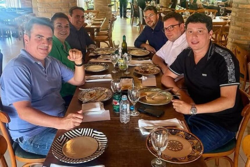 Seis homens sentados ao redor de uma mesa de madeira em restaurante com decoração rústica, incluindo pilares de tijolos e teto com estrutura de madeira. A mesa está posta com pratos, talheres, copos e garrafas de água. Ao fundo, outras pessoas estão em pé e sentadas, com luz natural entrando pelas janelas.