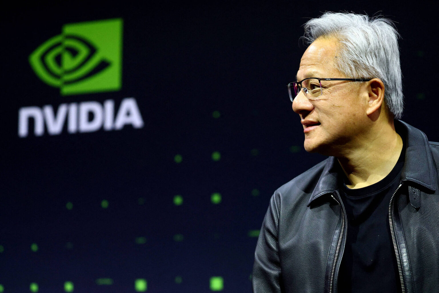 Nvidia: ações podem oscilar US$ 280 bilhões após balanço Homem de cabelos grisalhos e óculos, vestindo jaqueta preta, olha para a direita em frente a tela com o logotipo verde e branco da Nvidia.