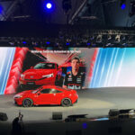 Carro esportivo vermelho exibido em palco com grande tela ao fundo mostrando imagem do veículo e piloto com uniforme Red Bull. Texto na tela convida para o Salão do Automóvel de São Paulo.