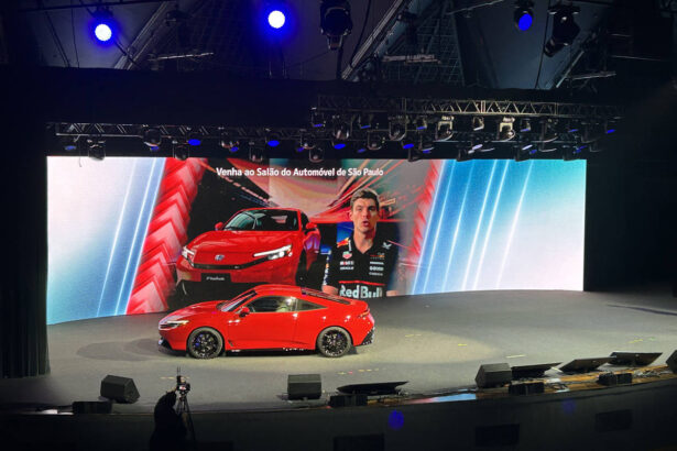 Carro esportivo vermelho exibido em palco com grande tela ao fundo mostrando imagem do veículo e piloto com uniforme Red Bull. Texto na tela convida para o Salão do Automóvel de São Paulo.