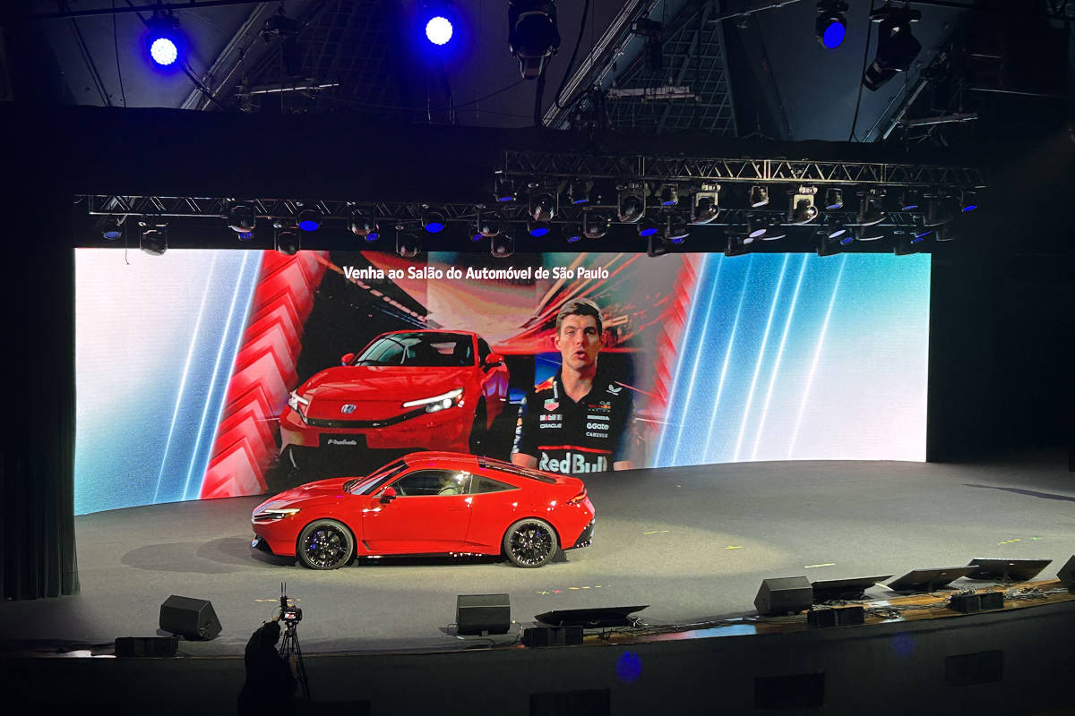 Carro esportivo vermelho exibido em palco com grande tela ao fundo mostrando imagem do veículo e piloto com uniforme Red Bull. Texto na tela convida para o Salão do Automóvel de São Paulo.
