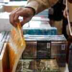 Mão de pessoa segurando e folheando um livro com capa amarela em mesa cheia de livros empilhados. Outra pessoa ao lado veste moletom bege com estampa colorida.