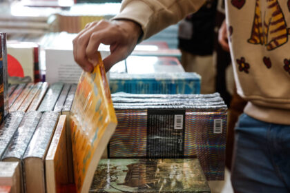 Mão de pessoa segurando e folheando um livro com capa amarela em mesa cheia de livros empilhados. Outra pessoa ao lado veste moletom bege com estampa colorida.