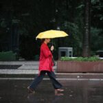 Pessoa vestindo casaco vermelho caminha na calçada molhada segurando um guarda-chuva amarelo aberto. Ambiente urbano com árvores e bancos ao fundo em dia escuro e chuvoso.