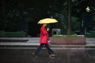 Pessoa vestindo casaco vermelho caminha na calçada molhada segurando um guarda-chuva amarelo aberto. Ambiente urbano com árvores e bancos ao fundo em dia escuro e chuvoso.