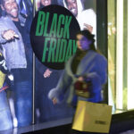 Pessoa com máscara facial caminha carregando sacola amarela em frente a vitrine iluminada com anúncio grande escrito