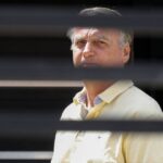 Homem de meia-idade com camisa polo amarela observa à frente, enquadrado por persianas horizontais pretas que criam faixas escuras na imagem.