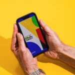 Bolsa Família: calendário de pagamento para dezembro de 2025 Mãos seguram um celular com tela exibindo formas geométricas coloridas em fundo amarelo. A sombra das mãos e do aparelho aparece à esquerda.