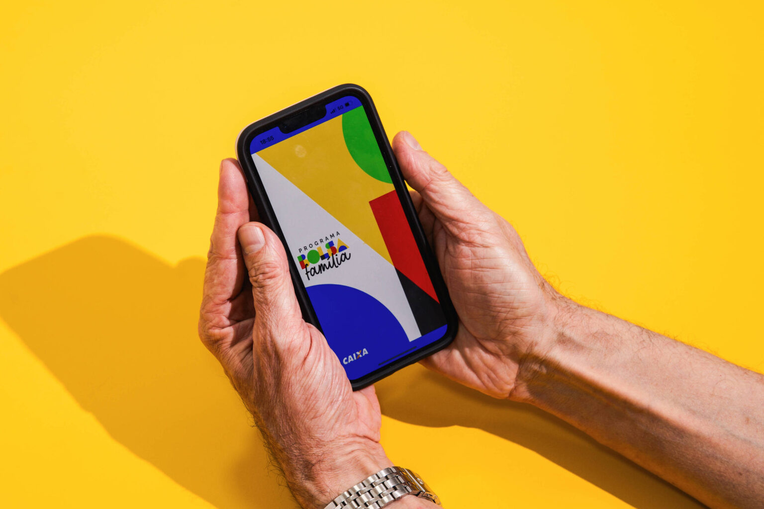 Bolsa Família: calendário de pagamento para dezembro de 2025 Mãos seguram um celular com tela exibindo formas geométricas coloridas em fundo amarelo. A sombra das mãos e do aparelho aparece à esquerda.