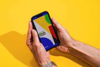 Mãos seguram um celular com tela exibindo formas geométricas coloridas em fundo amarelo. A sombra das mãos e do aparelho aparece à esquerda.
