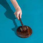 Justice do RJ mantém afastamento de diretoria da Oi até janeiro Mão segura martelo de juiz de madeira sobre base redonda de madeira, fundo azul.