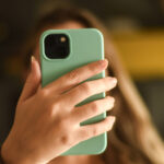 Febraban adverte sobre golpe de falso advogado aos consumidores A imagem mostra uma mão segurando um celular com capa verde. O fundo é desfocado, mas há uma superfície amarela visível. A mão parece feminina, com unhas bem cuidadas.