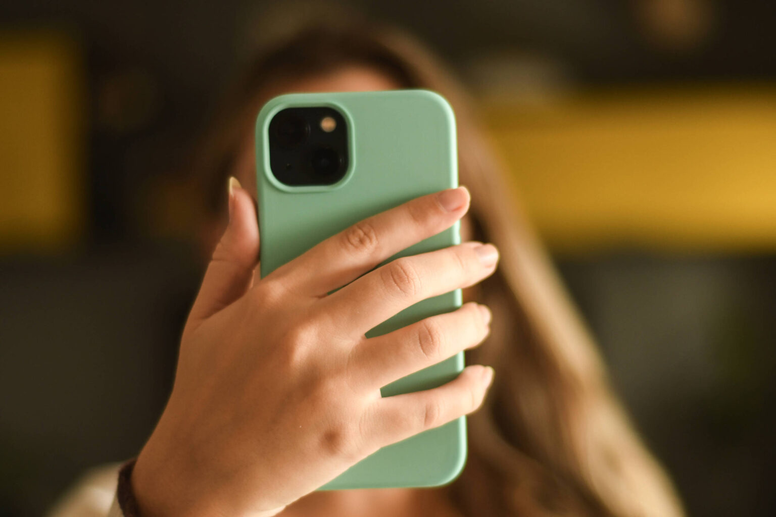 Febraban adverte sobre golpe de falso advogado aos consumidores A imagem mostra uma mão segurando um celular com capa verde. O fundo é desfocado, mas há uma superfície amarela visível. A mão parece feminina, com unhas bem cuidadas.