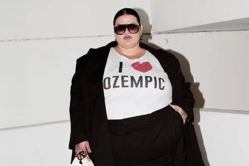 A imagem mostra uma mulher posando em um ambiente interno. Ela está vestindo uma camiseta branca com a frase