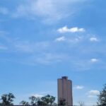 Céu azul claro com poucas nuvens brancas dispersas acima de telhados residenciais e árvores. Um prédio alto aparece ao fundo, sob luz do dia clara.