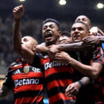 Quatro jogadores do Flamengo vestindo camisa preta e vermelha comemoram juntos com expressões intensas. Um deles está com o punho erguido, enquanto os outros o abraçam. O fundo mostra arquibancada desfocada e iluminação do estádio.