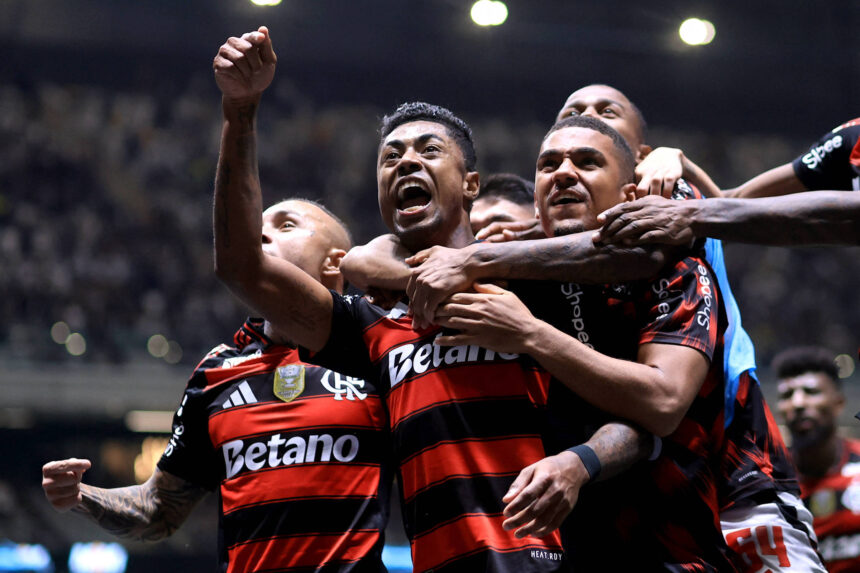 Quatro jogadores do Flamengo vestindo camisa preta e vermelha comemoram juntos com expressões intensas. Um deles está com o punho erguido, enquanto os outros o abraçam. O fundo mostra arquibancada desfocada e iluminação do estádio.