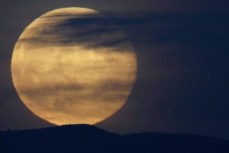 Lua cheia grande aparece parcialmente encoberta por nuvens finas e escuras, com silhueta de colina ao fundo em céu escuro.