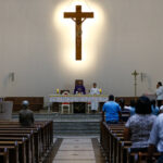 Interior de igreja com fiéis sentados em bancos de madeira voltados para altar. Padre e acólito estão no altar, com crucifixo iluminado na parede ao fundo. Ambiente claro e simples, com poucas pessoas presentes.