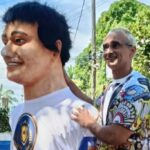 Carnavalesco de Olinda falece em academia Duas pessoas estão em área externa com árvores e céu azul ao fundo. A pessoa à frente usa camiseta branca com estampa colorida e sorri olhando para a esquerda. A pessoa atrás, com camiseta estampada colorida e shorts pretos, sorri enquanto toca nos ombros da pessoa à frente.