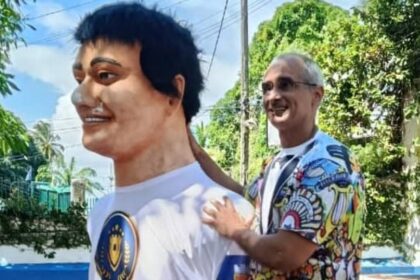 Duas pessoas estão em área externa com árvores e céu azul ao fundo. A pessoa à frente usa camiseta branca com estampa colorida e sorri olhando para a esquerda. A pessoa atrás, com camiseta estampada colorida e shorts pretos, sorri enquanto toca nos ombros da pessoa à frente.