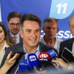 Político central fala à imprensa cercado por microfones de várias emissoras, com equipe ao fundo e banner azul com número 11.