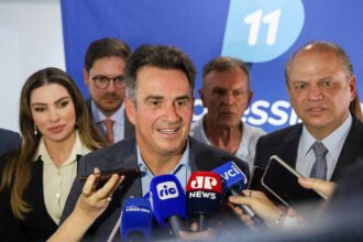 Político central fala à imprensa cercado por microfones de várias emissoras, com equipe ao fundo e banner azul com número 11.