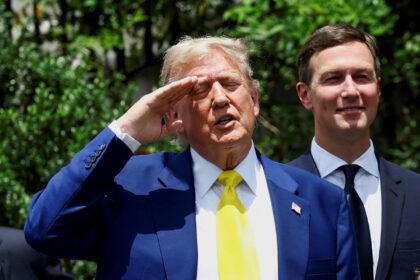 Warner Bros: Genro de Trump é envolvido em proposta de venda Ex-presidente dos Estados Unidos em terno azul e gravata amarela faz saudação com a mão direita na testa. Homem ao lado veste terno escuro e camisa branca, sorrindo. Fundo com vegetação e luz solar.