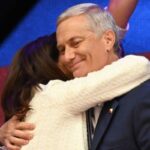 Chile: Kast é eleito e direita retorna ao poder em 2025 Homem de cabelos grisalhos veste terno azul escuro e sorri com os olhos fechados enquanto é abraçado por mulher de cabelos castanhos e casaco branco. Fundo desfocado com luzes coloridas.